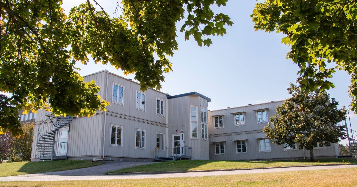 Flygfoto över ett skolområde omgivet av skog och en sjö, där traditionella skolbyggnader med röda tak syns bredvid grå modulbyggnader och en stor lekplats med barn,